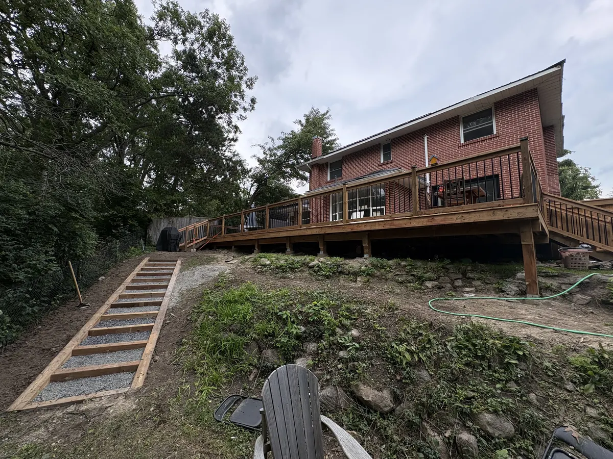 Raised wood deck with black baluster railing and stairs behind a brick house on a sloped back yard